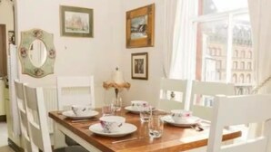 Un coin repas avec une table en bois dressée pour quatre personnes, des chaises blanches et des tableaux encadrés accrochés au mur.