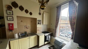 Une cuisine équipée d’une cuisinière à gaz, d’un évier et de placards. Il y a une fenêtre avec des rideaux et une vue sur la rue.