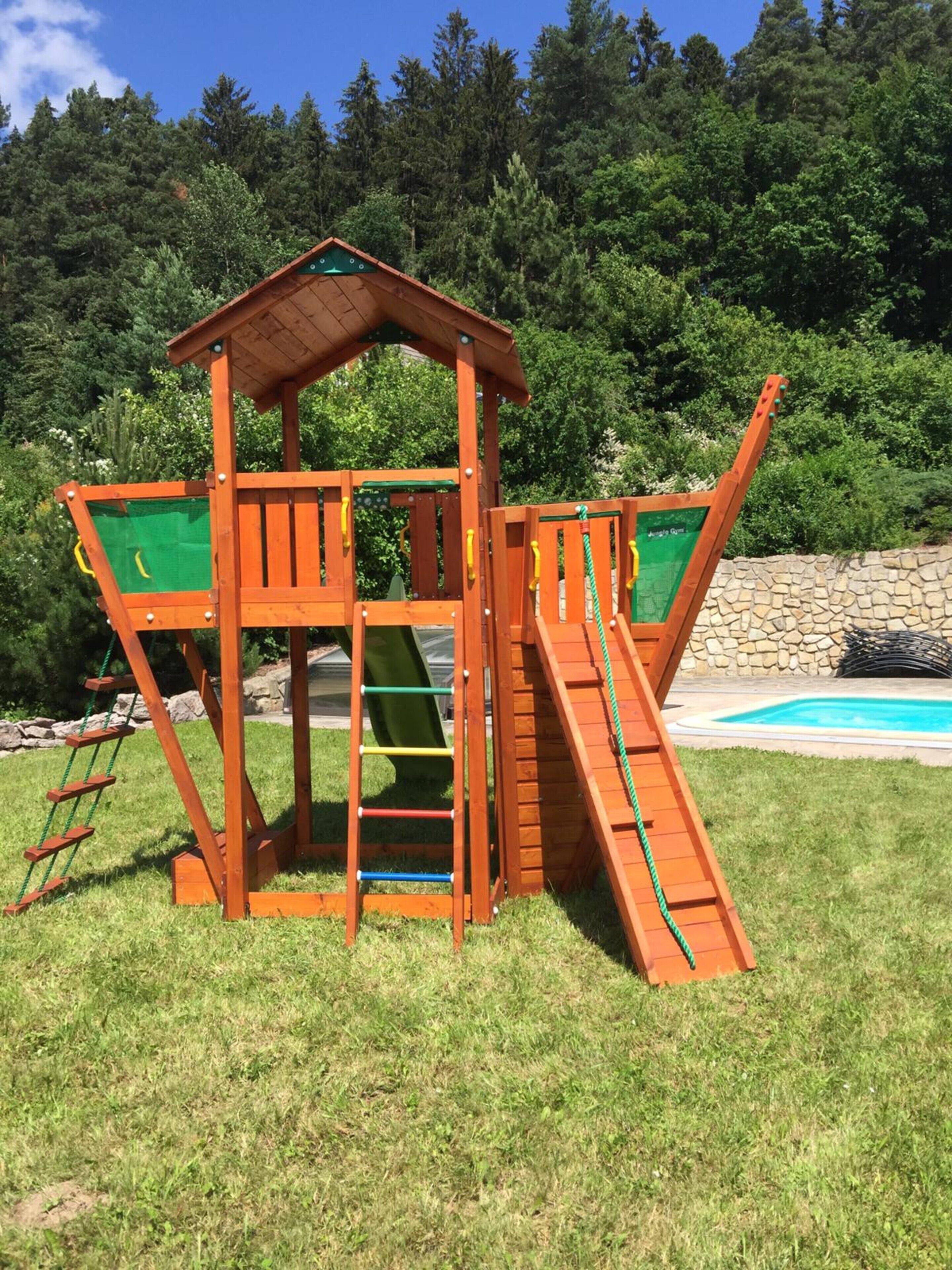 A wooden play structure with a slide and climbing elements, set in a grassy area with a pool and trees in the background.