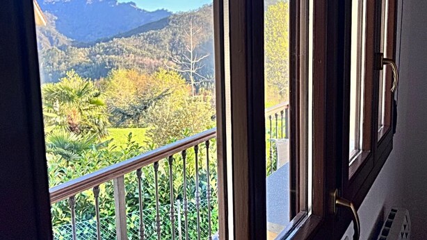Blick aus einem Fenster mit Holzrahmen und Metallgitter auf eine gebirgige Landschaft mit Bäumen und blauem Himmel.