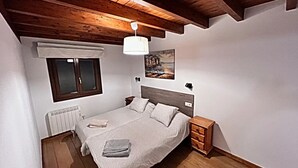 Ein Schlafzimmer mit Holzbalkendecke, einem Bett mit weißem Kopfteil, einem Nachttisch, einem Fenster mit Jalousien und einem Heizkörper.