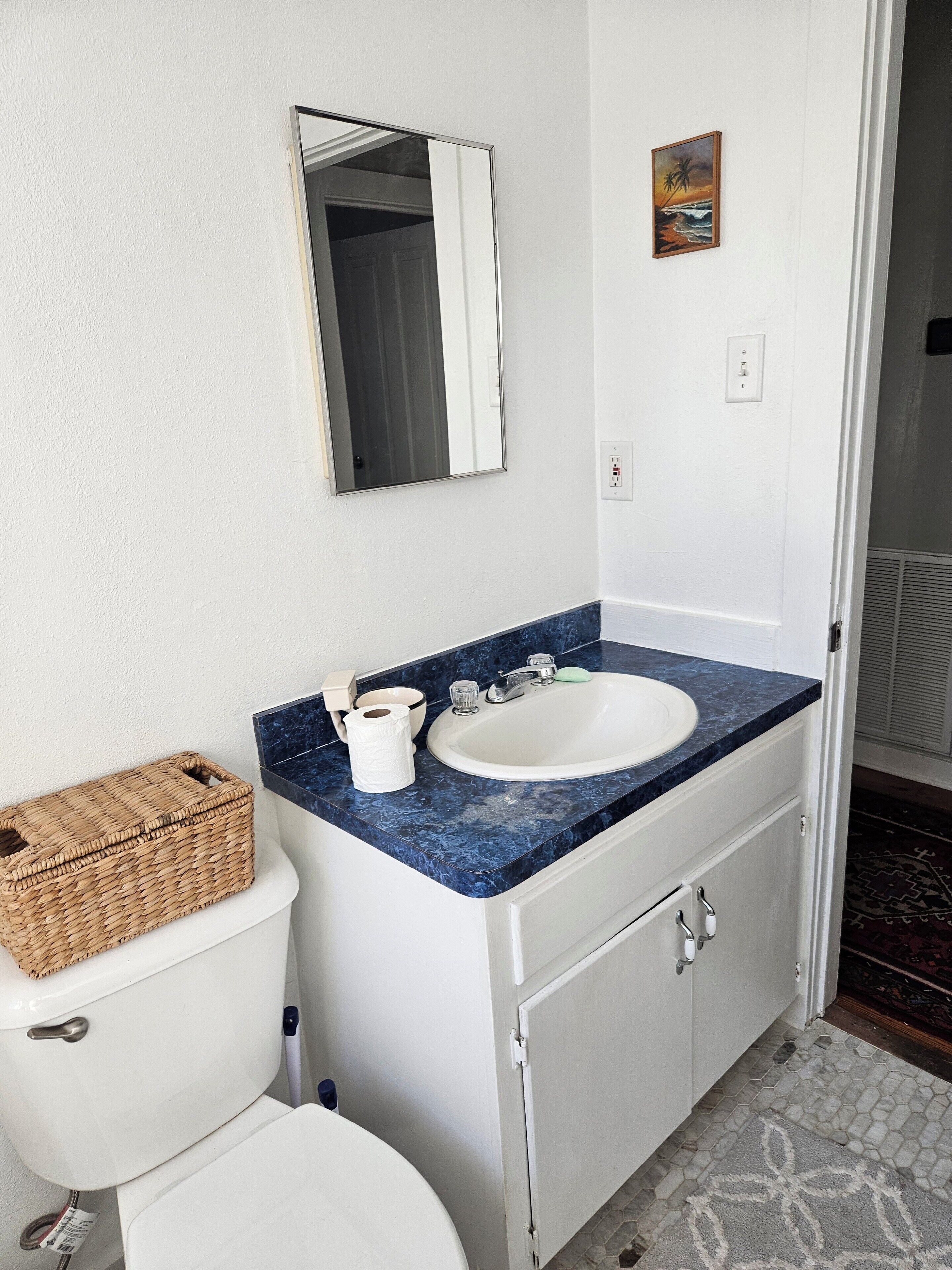 Un bany amb un vàter blanc, un lavabo amb taulell de marbre blau, un mirall i un petit quadre a la paret.