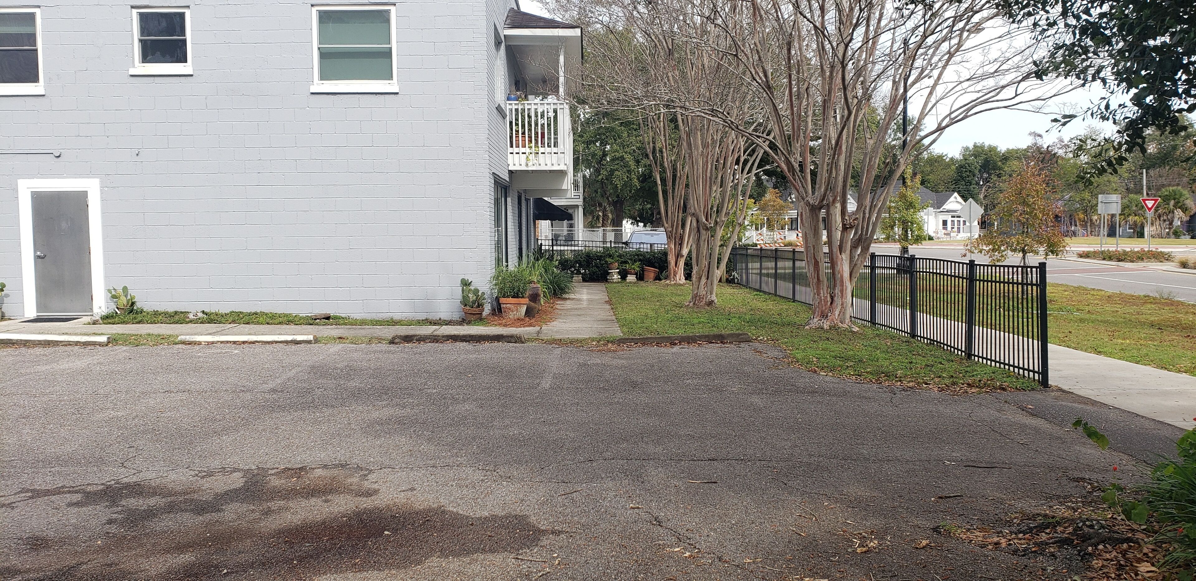 A residential street with a grey apartment building, a black metal fence, and a sidewalk.
