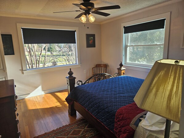 A bedroom with a bed, a ceiling fan, two windows with blinds, a wooden floor, and a nightstand with a lamp.