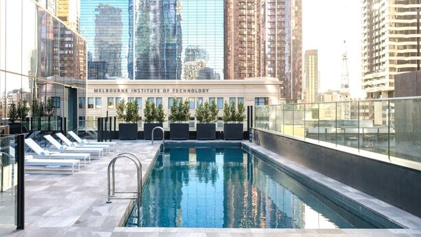 Egy tetőtéri úszómedence, napozóágyakkal, üvegkorláttal, a háttérben pedig a Melbourne-i Műszaki Intézet épülete látható.