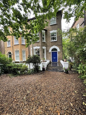 A multi-story brick building with a blue door, surrounded by trees and a gravel driveway.