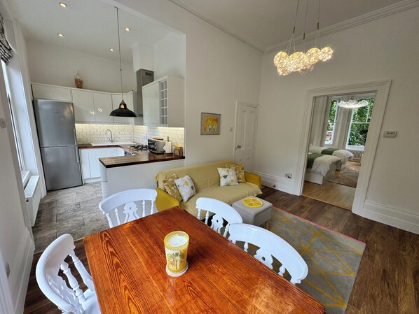 A modern kitchen with a refrigerator, stove, and white cabinets. There is a dining area with a wooden table and white chairs. A sofa is visible in the background.