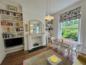 A room with a fireplace, bookshelves, a dining table, and a large window with a view of greenery.