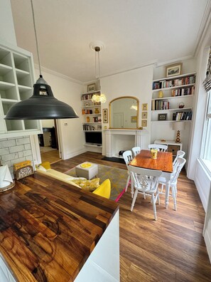 A modern kitchen with a wooden island, white chairs, a dining table, and a bookshelf filled with books.