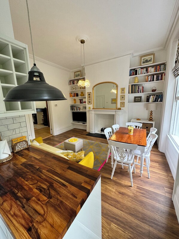 A modern kitchen with a wooden island, white chairs, a dining table, and a bookshelf filled with books.