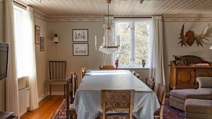 Een eetkamer met een lange tafel, stoelen, een kroonluchter, een radiator en een raam met gordijnen.