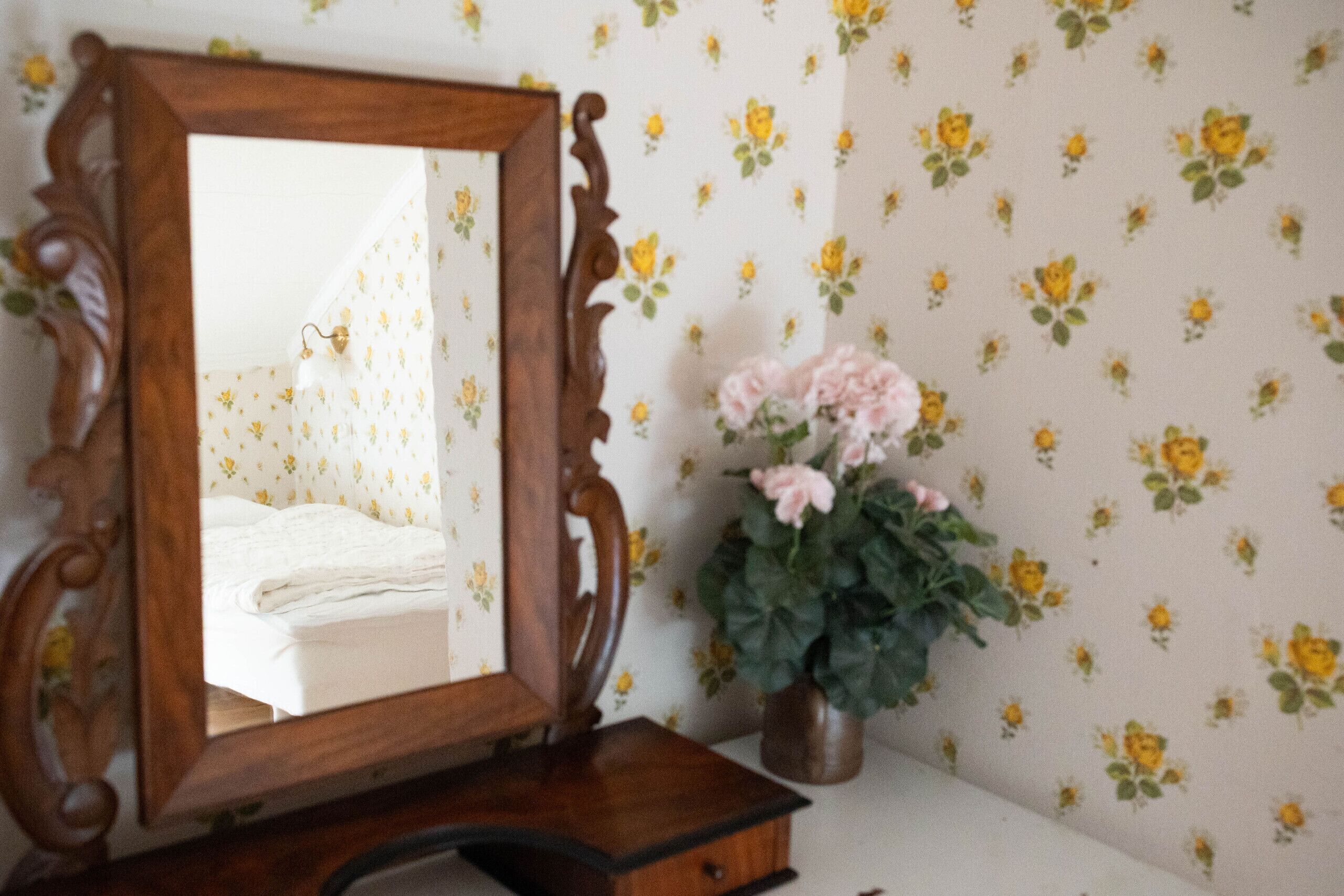 A wooden dresser with a mirror, a small vase with pink flowers, and a floral wallpaper pattern.