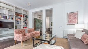 A modern living room with a flat-screen TV, a glass coffee table, patterned sofas, a bookshelf, and a dining area.