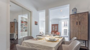 A dining area with a table set for four, surrounded by wicker chairs. There are bookshelves and a wooden cabinet in the background.