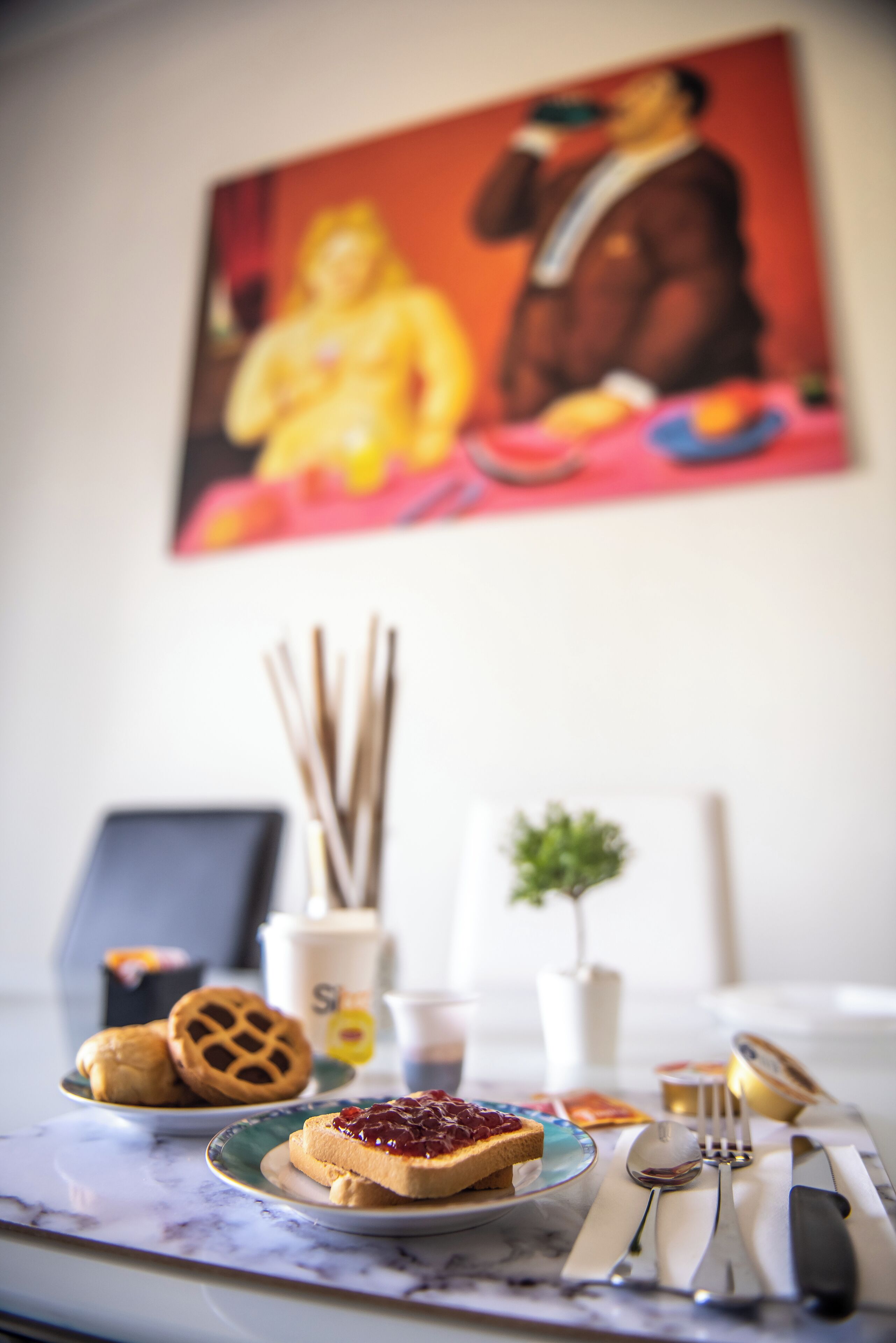 Una mesa de desayuno con tostadas y mermelada, una planta pequeña y un cuadro en la pared.