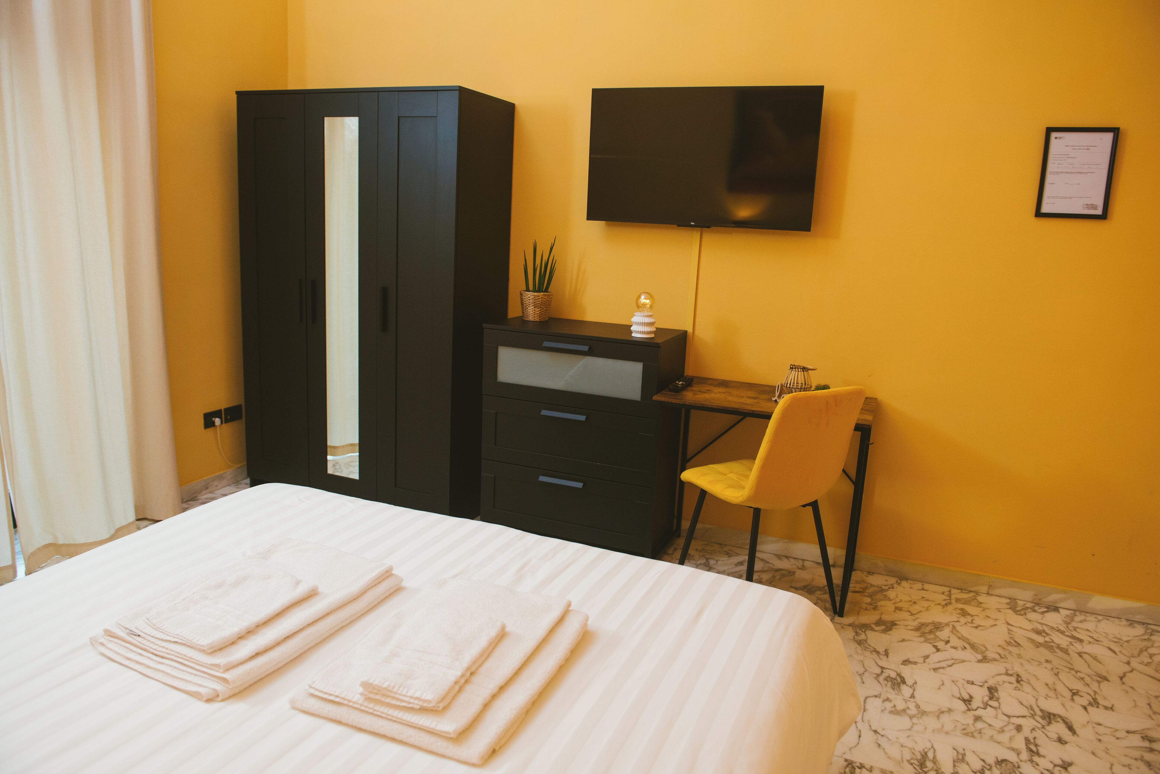 A hotel room with a bed, a wardrobe, a desk with a chair, a television, and a small plant.