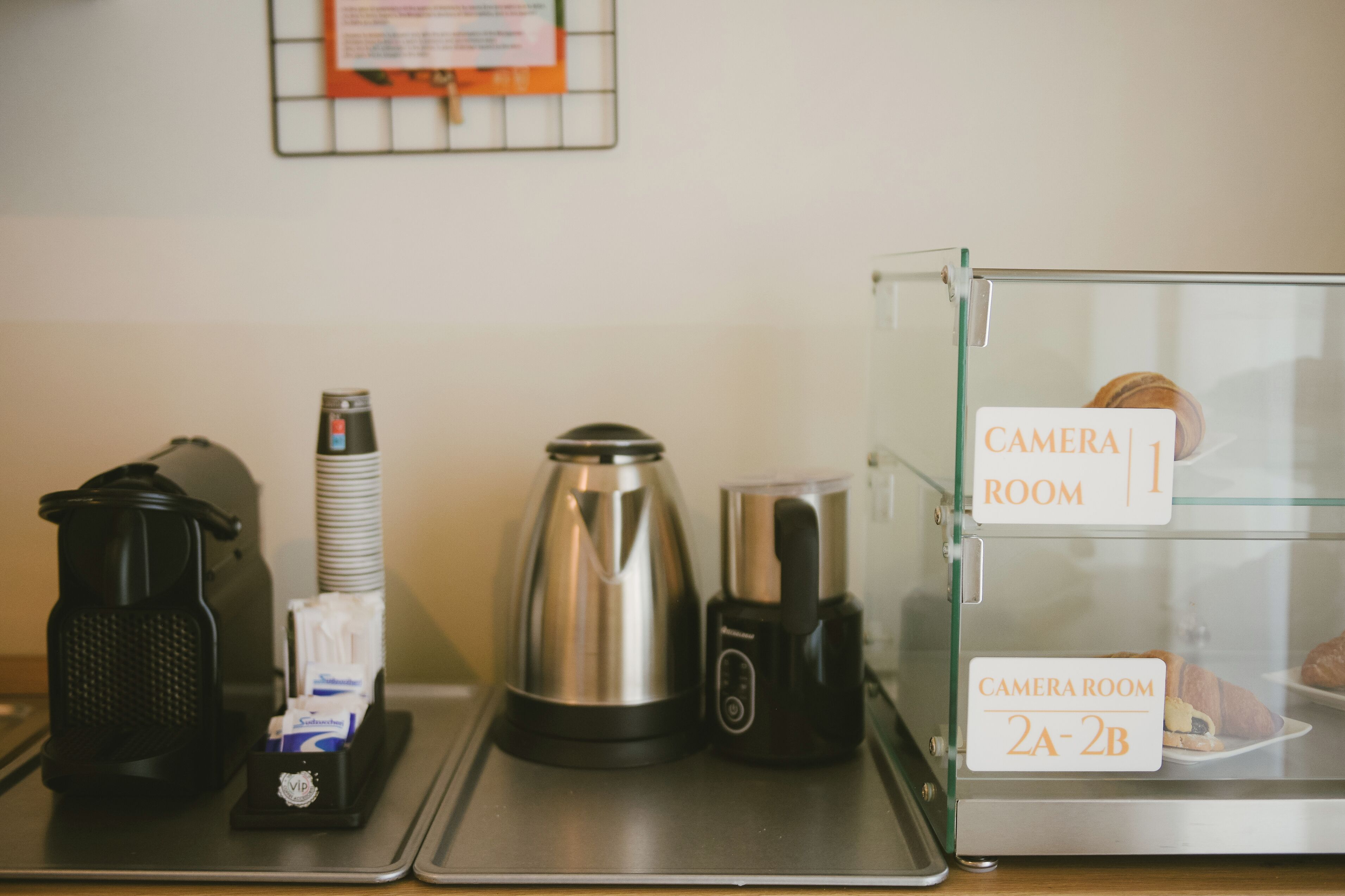 A coffee station with a coffee maker, a grinder, and a coffee pot on a tray. There is a glass display case labeled "Camera Room" with compartments marked "2A-2B".