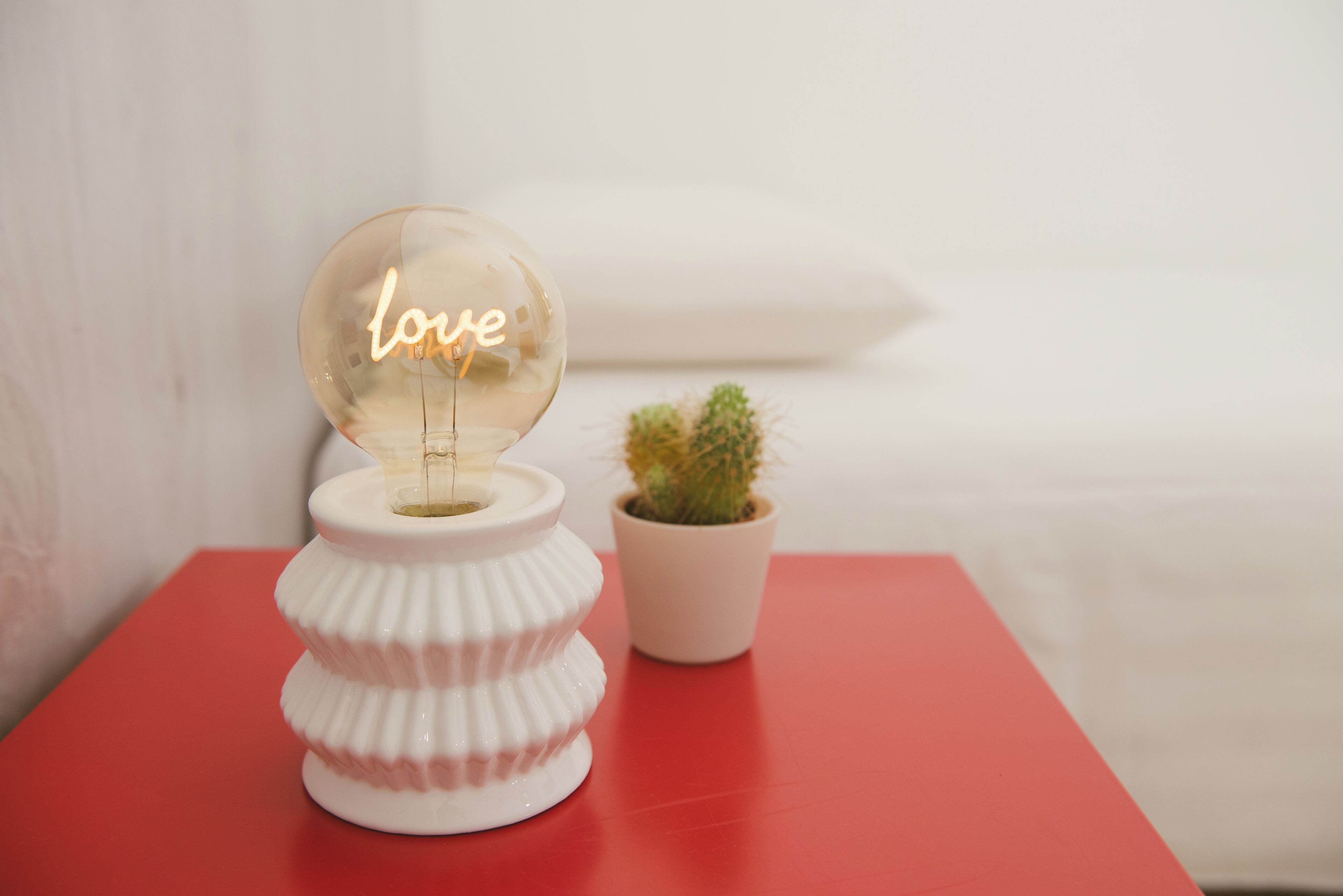 Un globo di vetro trasparente con la scritta 'love' all'interno, appoggiato su un piedistallo bianco accanto a un piccolo cactus in vaso su un tavolo rosso.