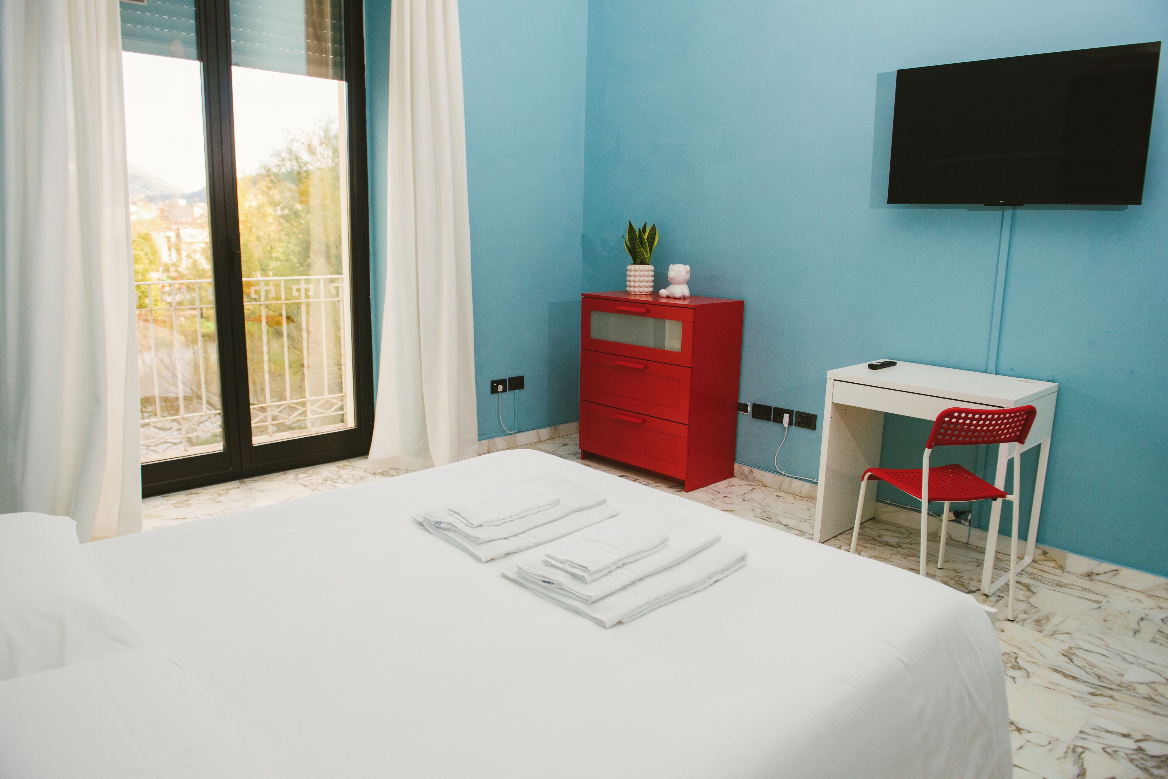 A hotel room with a bed, a desk, a red dresser, a TV, and a balcony with a view.