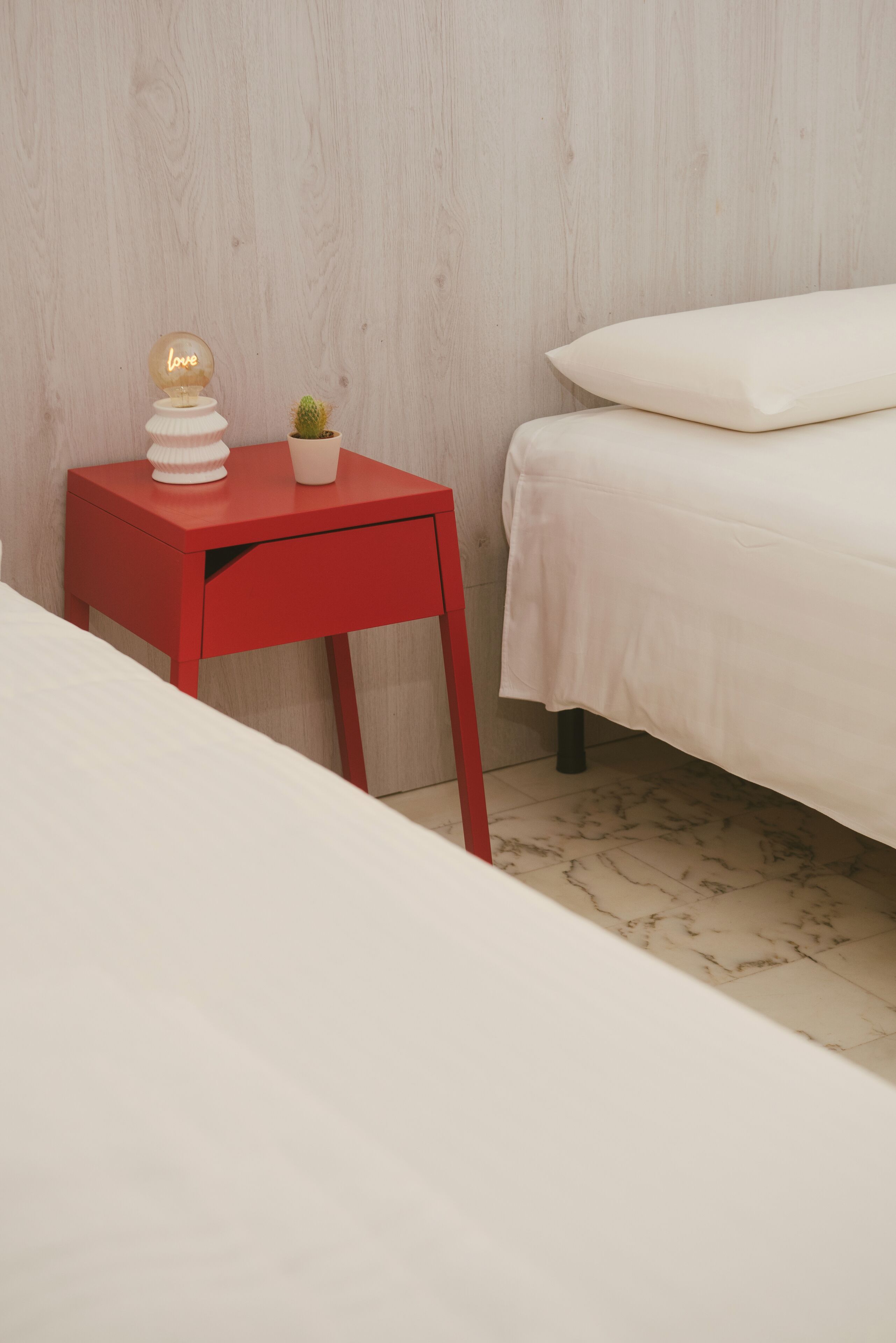 Une chambre avec une table de chevet rouge, un lit blanc et une petite plante en pot.