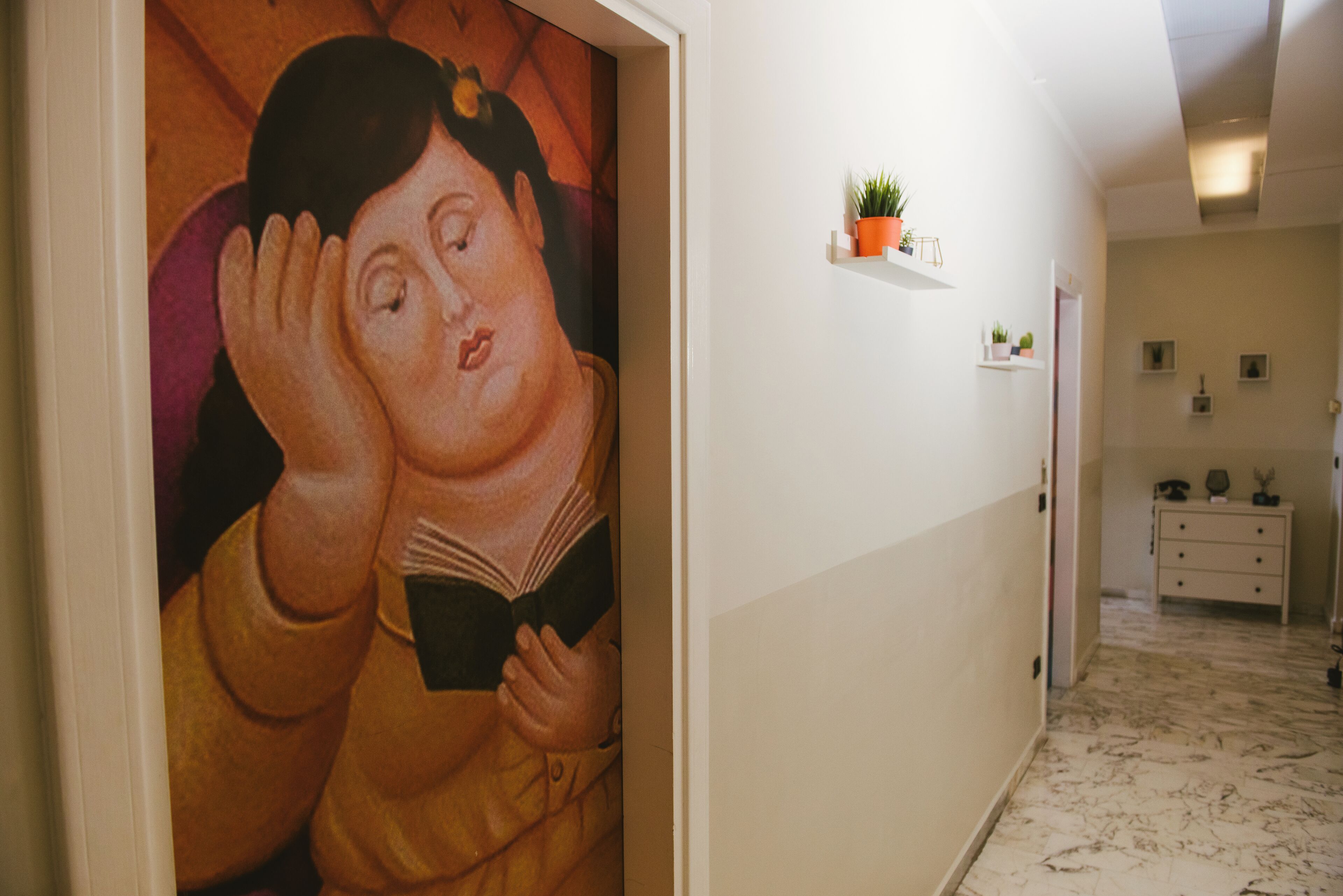 A hallway with a mural of a person reading a book, a white wall with a shelf holding a plant, and a marble floor.
