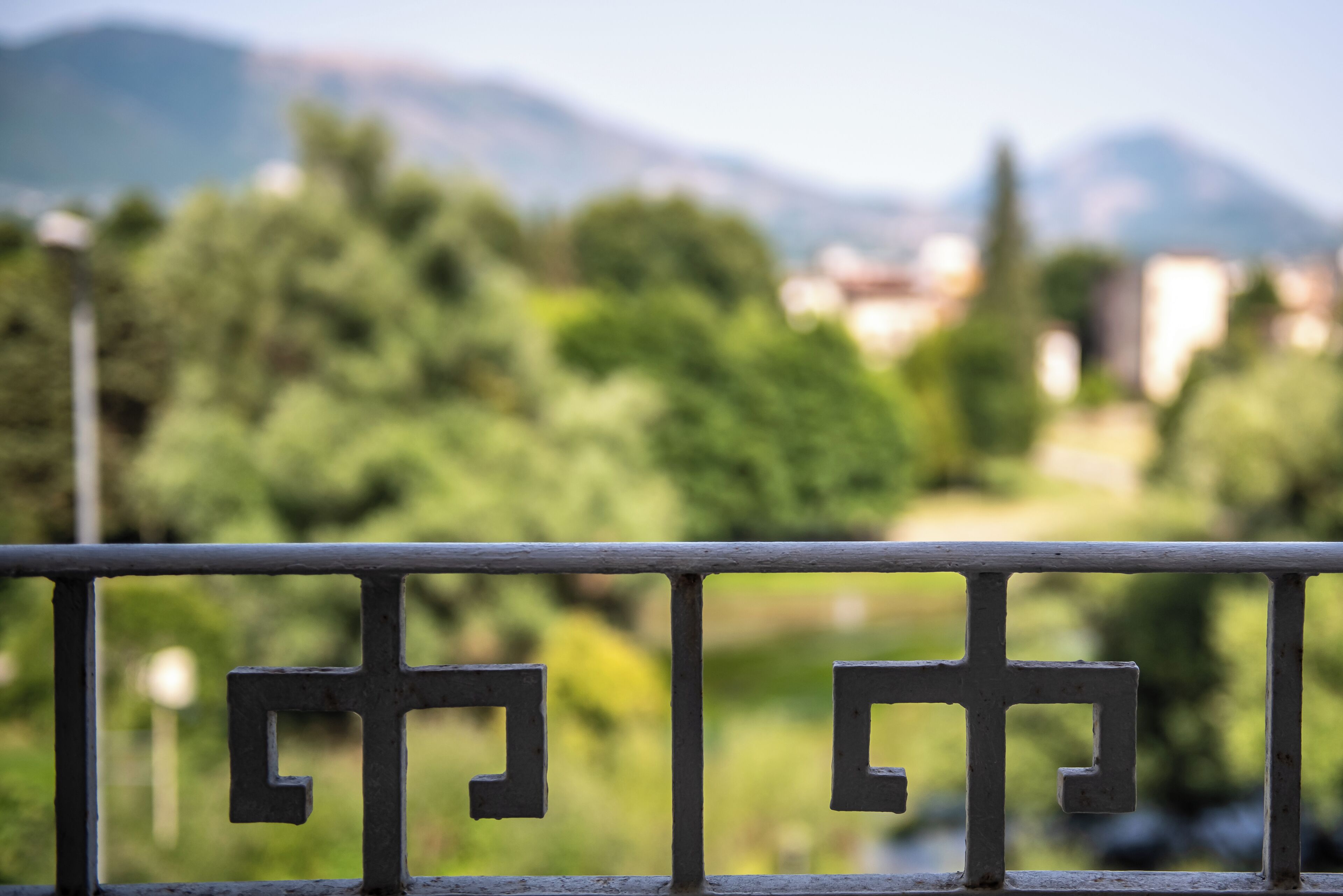 A metal railing with a geometric pattern, overlooking a river with trees and mountains in the background.