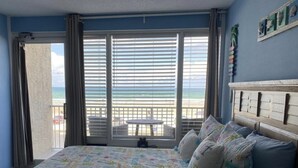 Une chambre à coucher avec un lit, des oreillers, une vue sur la plage et un balcon équipé d’une table et de chaises.
