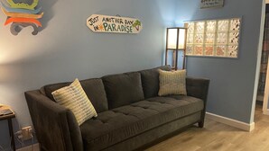Un salon avec un canapé marron foncé, deux coussins décoratifs à motifs, un panneau mural portant l’inscription « Encore une journée au paradis », et un élément décoratif mural sur le thème de la plage.