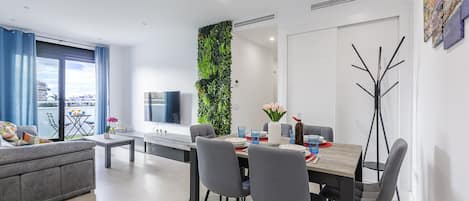 A modern living room with a grey sofa, a dining table with chairs, a TV, and a green vertical garden.