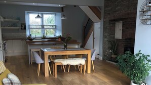 A spacious living area with a wooden dining table, yellow sofa, and a grey sofa. A kitchen with a stone wall and a staircase is visible in the background. - Renovated farmhouse, 5 mins from the sea (Ferwig)