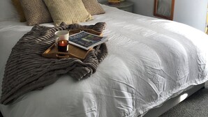 A neatly made bed with a white comforter, a brown throw blanket, and a tray with a candle and a magazine. There is a vase with dried flowers on a nightstand beside the bed. - Renovated farmhouse, 5 mins from the sea (Ferwig)