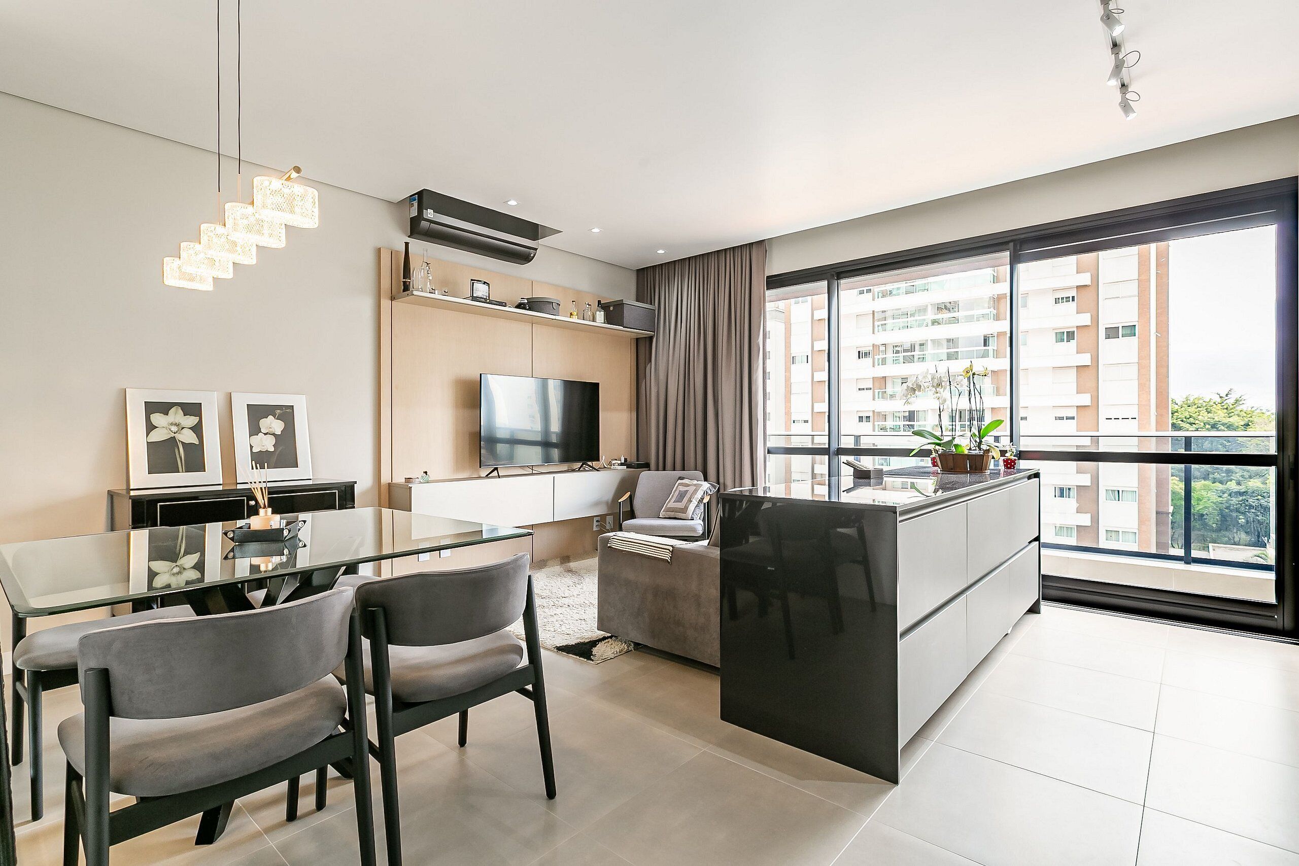 A modern living room with a glass dining table, grey chairs, a flat-screen TV, and a large window with a city view.