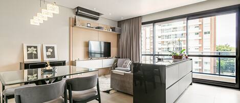 A modern living room with a glass dining table, grey chairs, a flat-screen TV, and a large window with a city view.