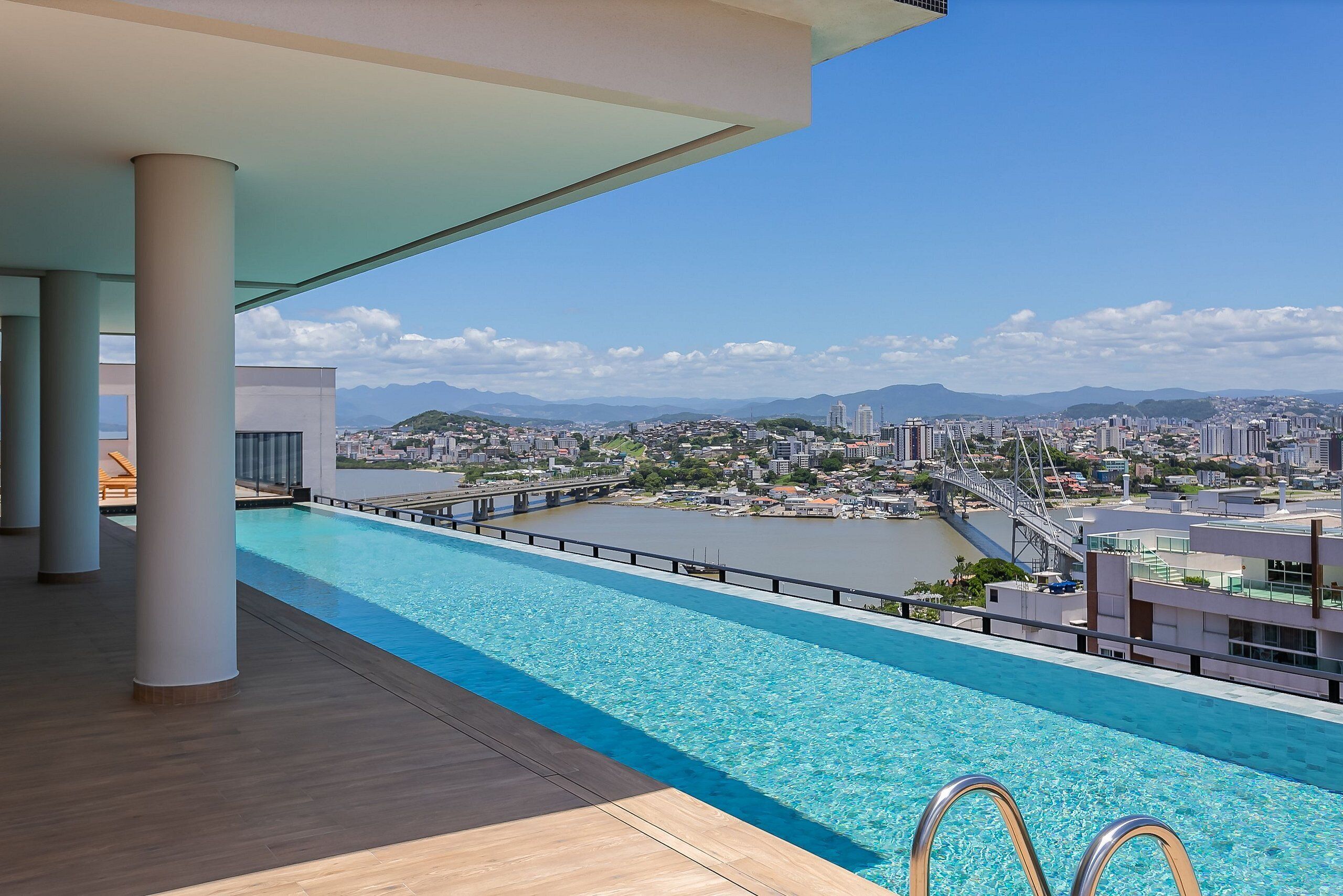 A rooftop infinity pool with a city view, large pillars, and a wooden deck.