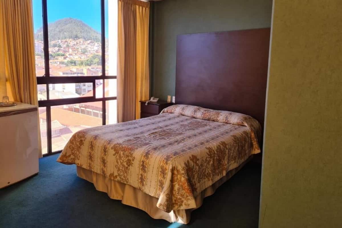 Quarto de hotel com cama, escrivaninha e vista para uma montanha e cidade através de grandes janelas.