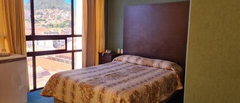 A hotel room with a bed, a desk, and a view of a mountain and town through large windows.