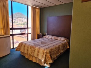 A hotel room with a bed, a desk, and a view of a mountain and town through large windows. - H. Glorieta (Sucre)