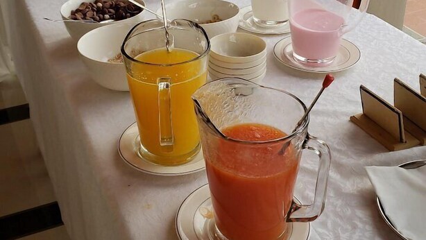 Tavolo per colazione a buffet con diversi cibi e bevande, tra cui una caraffa di succo d'arancia e un bicchiere di bevanda rosa, sullo sfondo di un cartello di un hotel.