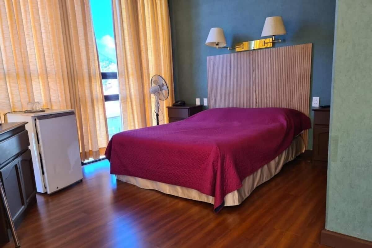 Quarto com cama, escrivaninha, ventilador e janela com cortinas.