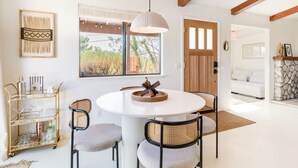 Un coin repas avec une table ronde, quatre chaises, un chariot doré contenant des verres, un luminaire suspendu et une fenêtre donnant sur des arbres.