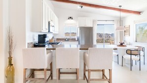 Une cuisine moderne avec des meubles blancs, un îlot central doté de tabourets de bar, un coin repas offrant une vue sur les montagnes et un luminaire suspendu.