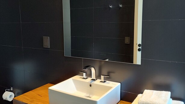 A modern bathroom with a white sink, two faucets, a mirror above, and a wooden vanity. - Main By Babel (Mendoza)