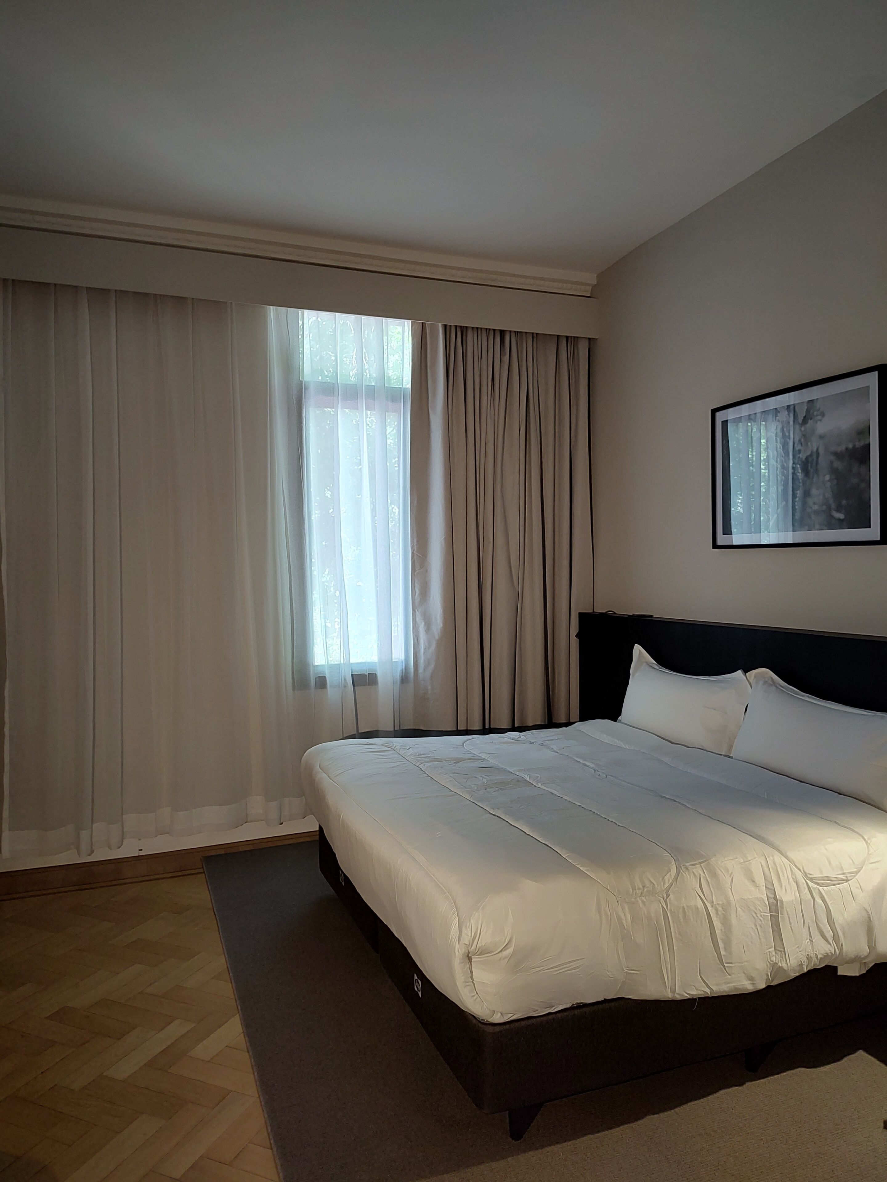 A hotel room with a large bed, a window with sheer curtains, and a framed picture on the wall.