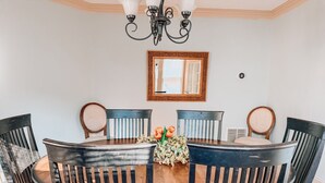A dining area with a round table, six chairs, a chandelier, a mirror, and a floral centerpiece.