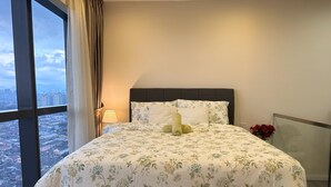 Habitación de hotel con una cama grande, ropa de cama floral, una mesita de noche con lámpara, un jarrón con flores rojas y vistas a un paisaje urbano a través de un ventanal amplio.
