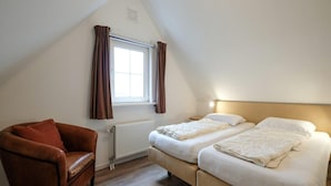 A small, cozy room with a sloped ceiling, a single window with curtains, a brown leather chair, and two beds with white bedding. - Holiday Home Near Beach and Forest (Sint Maartensvlotbrug)