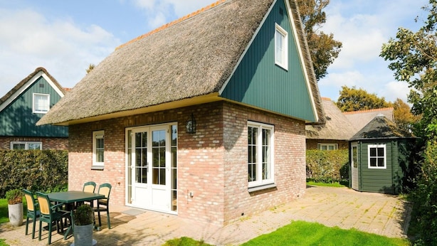 A single-story house with a thatched roof, brick walls, and a green exterior. It features a front door with glass panels, a small attached shed, and a paved patio with a table and chairs. - Holiday Home Near Beach and Forest (Sint Maartensvlotbrug)