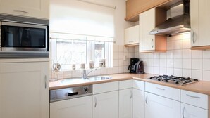 A modern kitchen with white cabinets, a stainless steel stove, a built-in oven, and a window with blinds. - Holiday Home Near Beach and Forest (Sint Maartensvlotbrug)