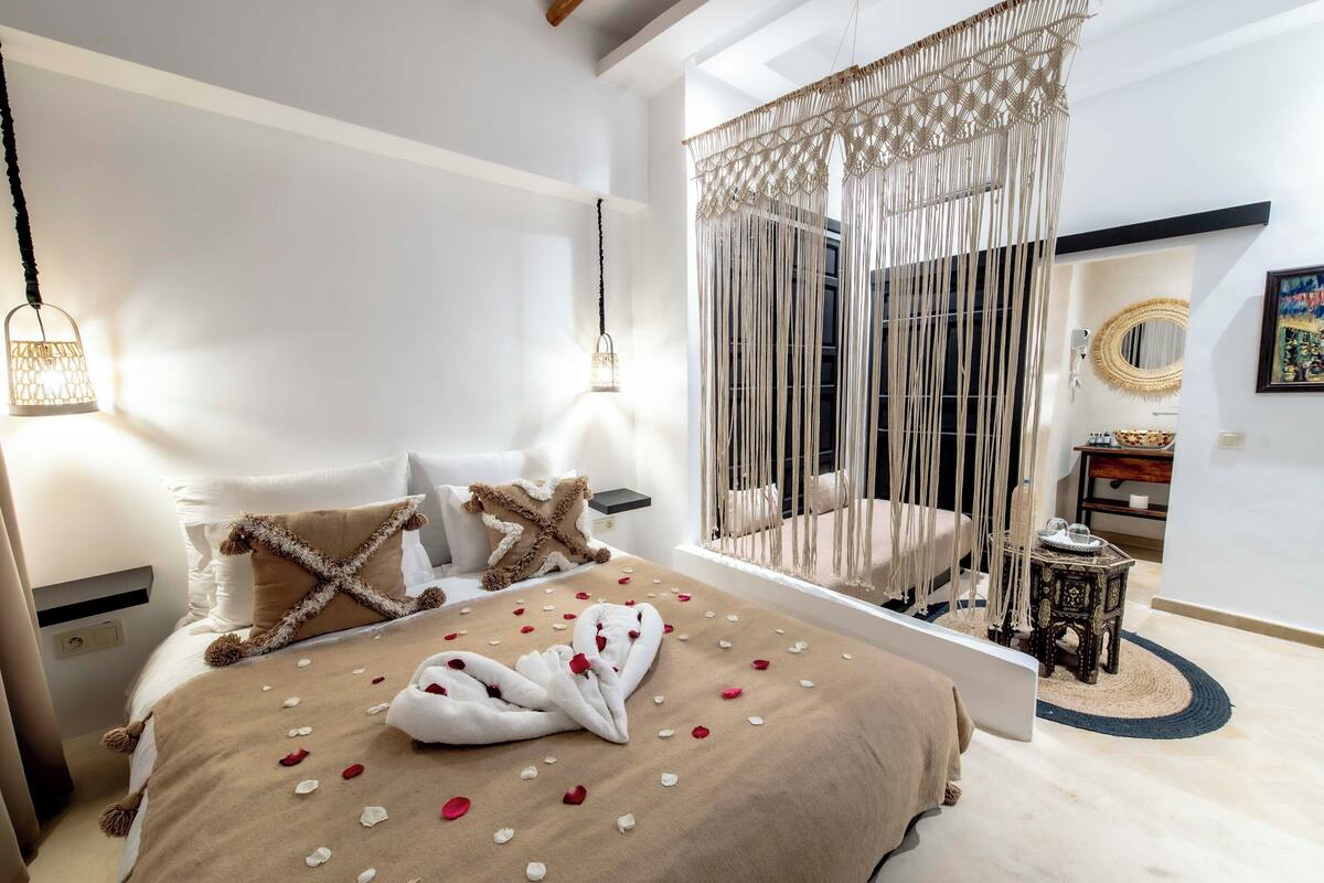 Une chambre bien éclairée, avec un lit, des lampes de chevet, un baldaquin en macramé et une petite table ronde surmontée d’un miroir.
