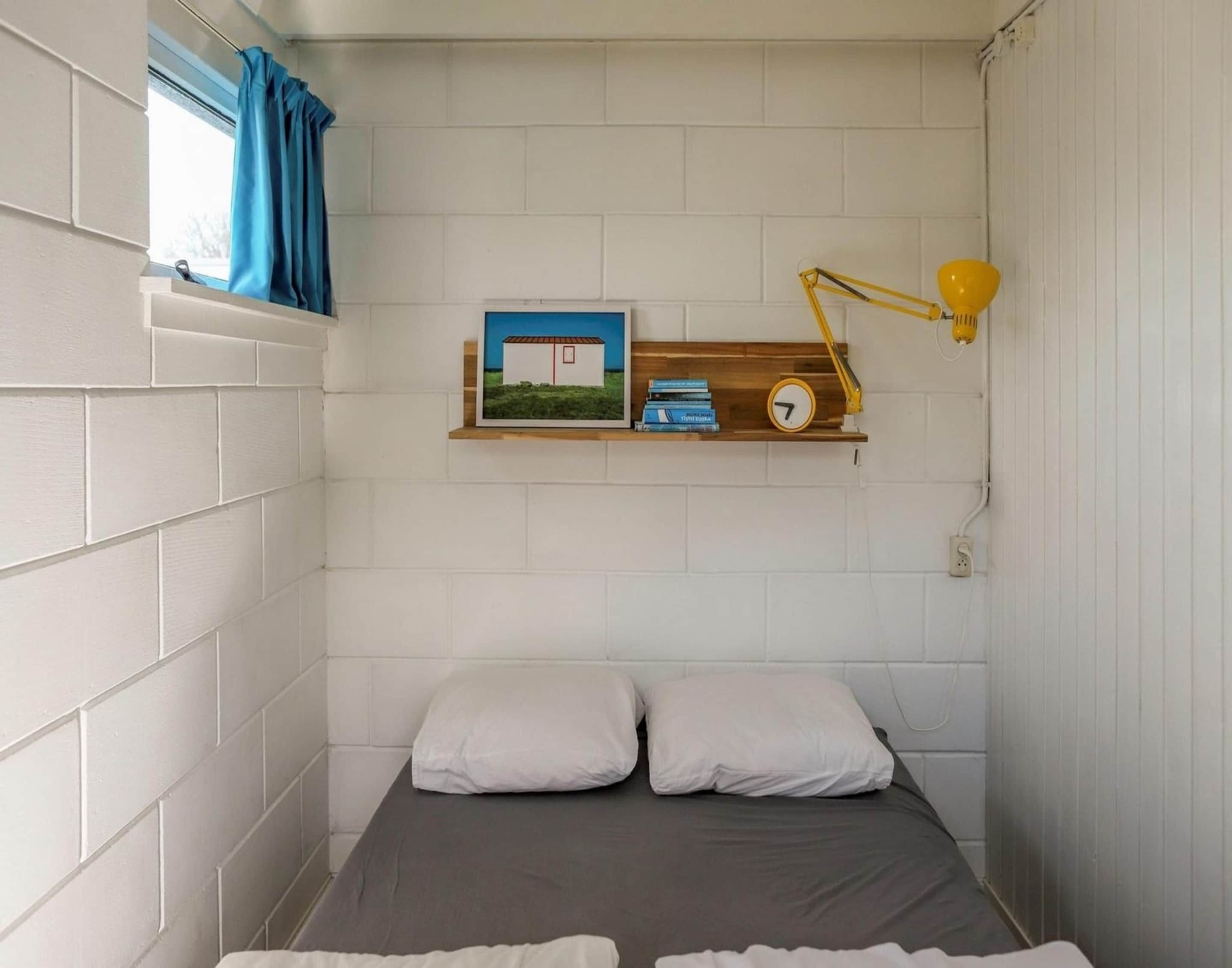 A small, compact bedroom with a bed, two pillows, a shelf with a clock and books, and a window with curtains.